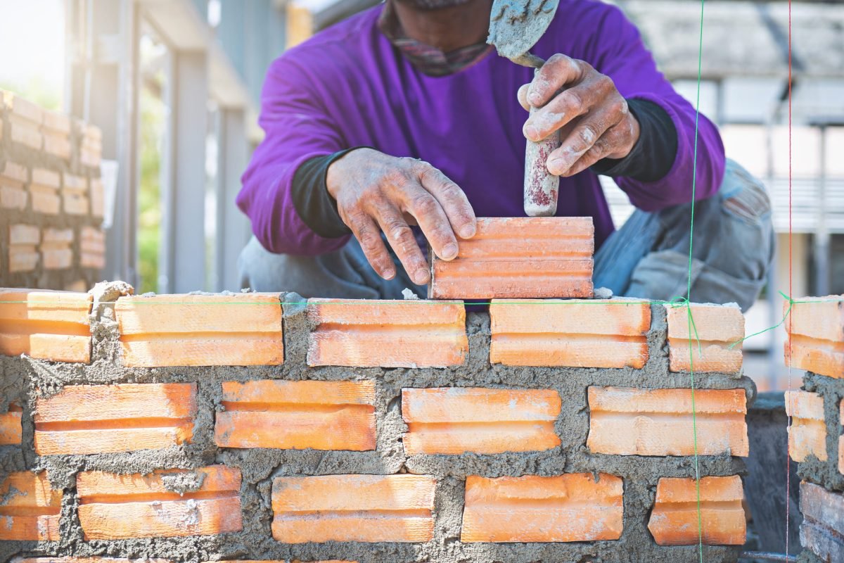 Bricklayer worker installing brick masonry on exterior wall with trowel putty knife for new house building at construction site.