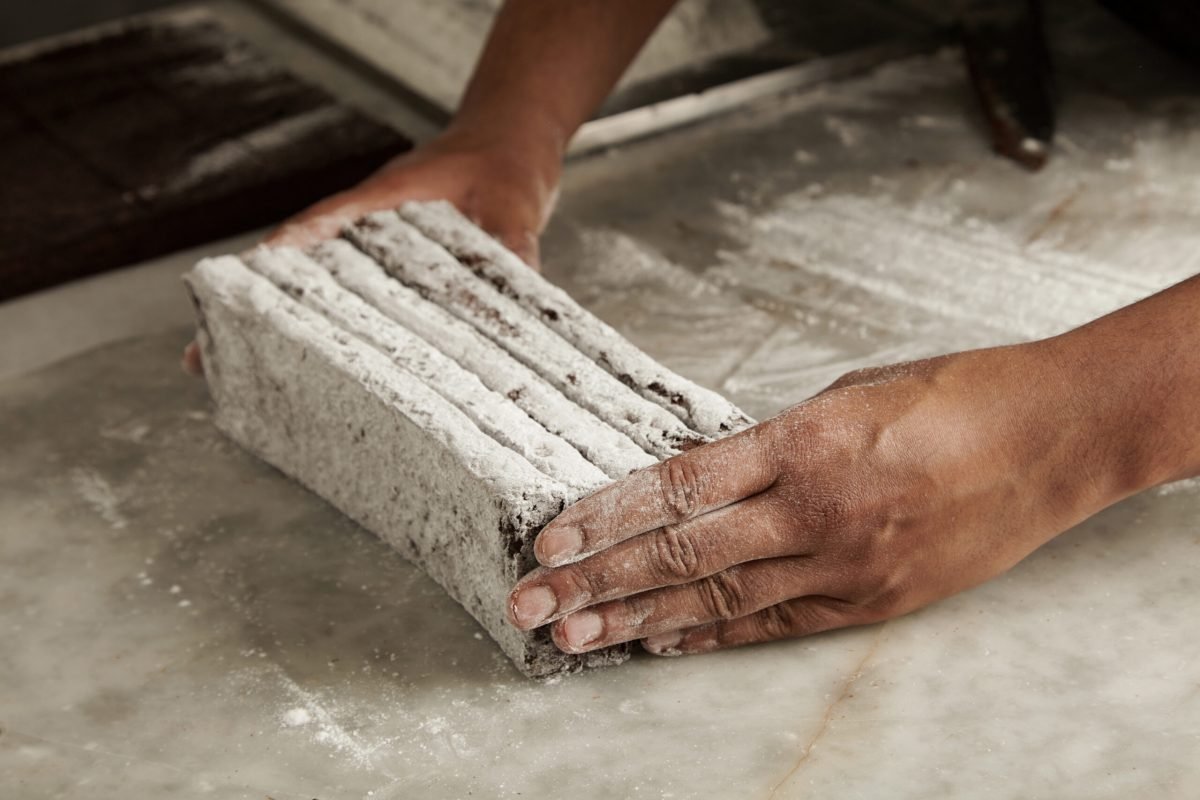 Hands of black man chief holds freshly baked chocolate bars in sugar powder before packing, closeup in professional artisan confectionery