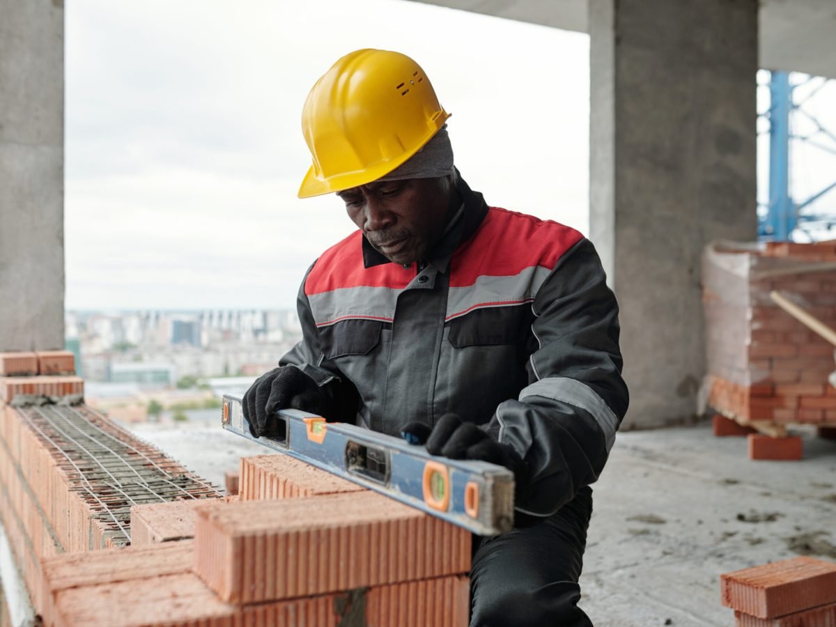 Mature builder in workwear and protective helmet keeping level handtool on surgace of brick layer while checking flatness of wall